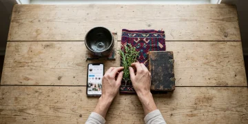 An overhead view of diverse cultural items, including a Japanese tea bowl, a smartphone, a Mexican textile, and a leather book, arranged on a wooden table. A hand places a green herb sprig amidst the artifacts, symbolizing hybrid cultural identities.