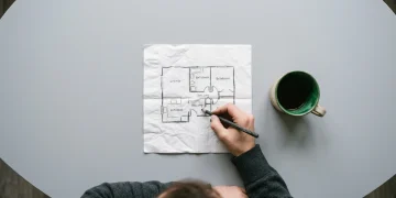 Overhead shot of a person sketching a room layout on a crumpled napkin with a charcoal pencil, a green ceramic mug beside it, illustrating the humble beginnings of a tech innovation.