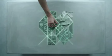 An overhead view of a hand arranging glowing green geometric shapes representing diverse cultural patterns like African mudcloth and Korean hanbok, on a concrete slab, symbolizing global cultural fusion and identity shifts.